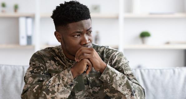 Military veteran sitting on a couch with hands folded near chin, as if staring in concern.